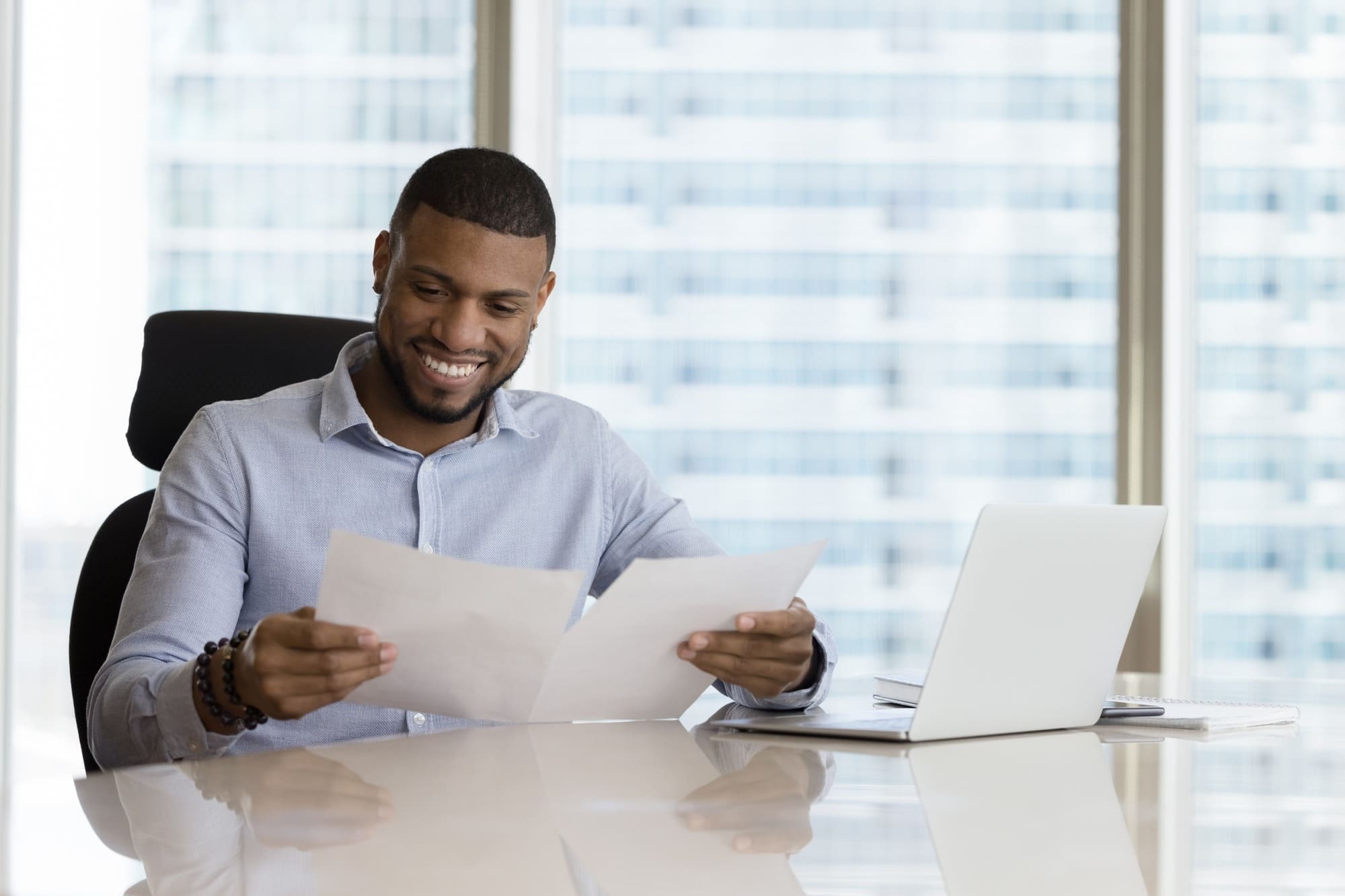 A smiling man reads a document while sitting in front of a laptop